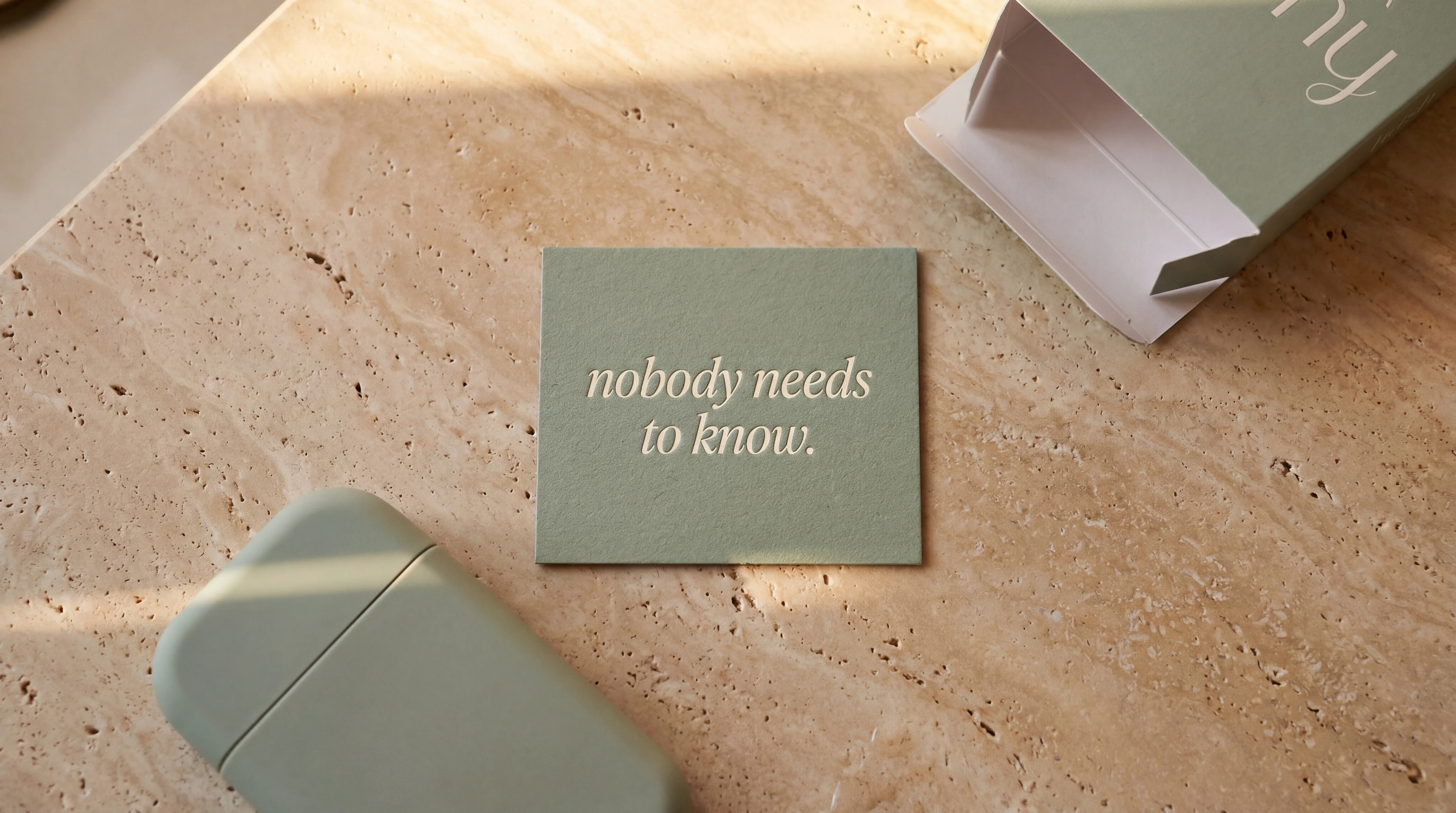 A small sage card printed with 'nobody needs to know.' in italic cream serif, resting on a warm travertine counter between a Hush bottle and box